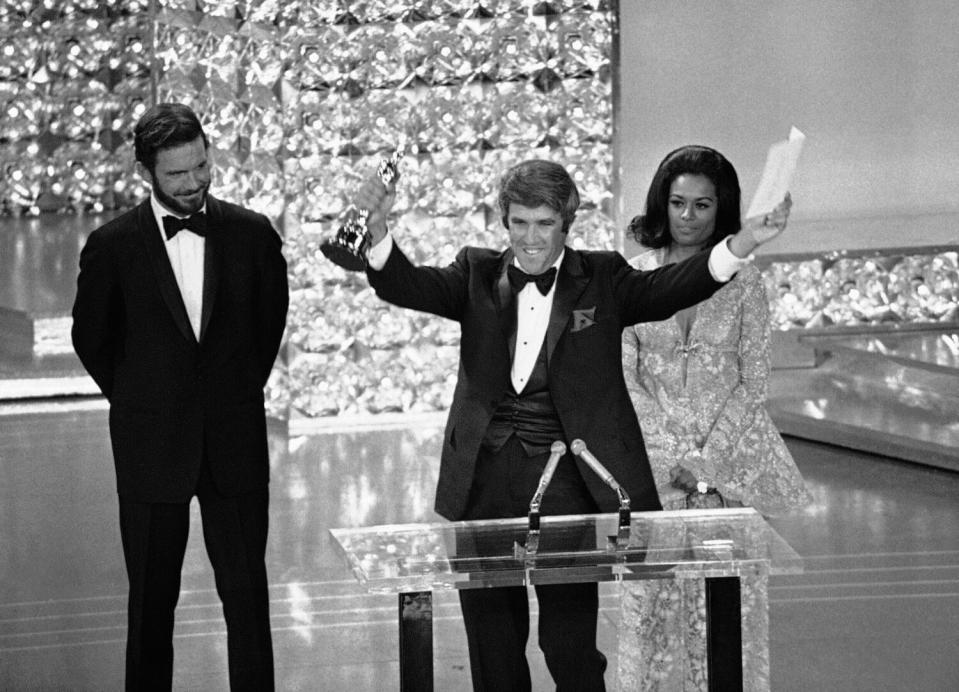 A man onstage holds out a trophy with his arms wide open