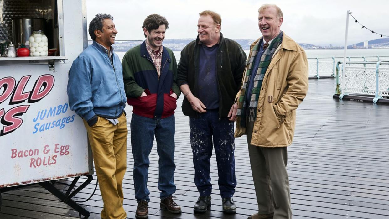  The cast of Men Up stand on a wet and rainy seaside pier in Wales next to a hut selling "jumbo sausages" (geddit?) . 