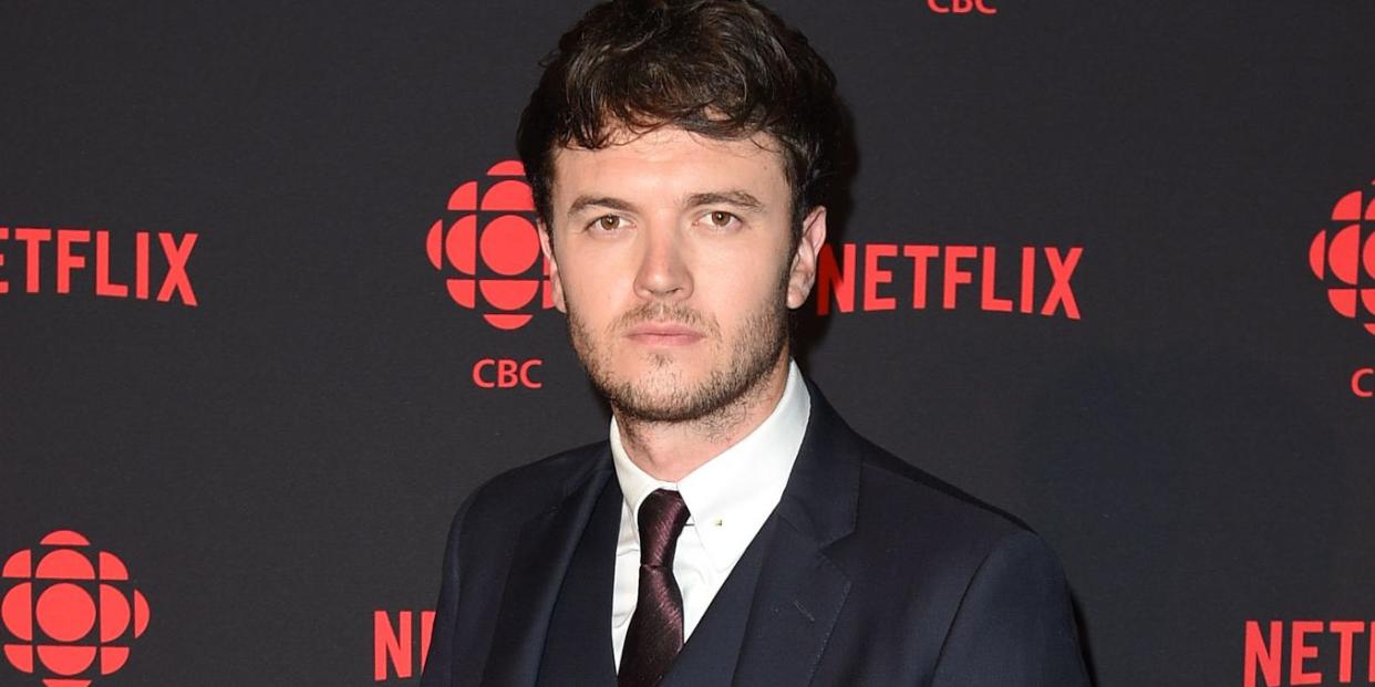 kerr logan, a man stands looking at the camera with a neutral facial expression, he has brown hair and a beard, he wears a navy suit with brown shoes