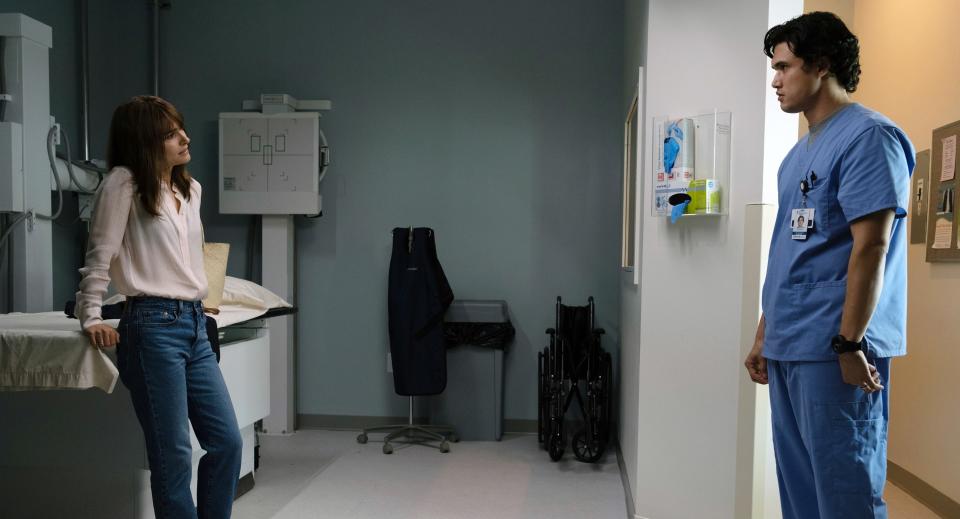 Joe in scrubs as he stands in the doorway of a hospital room looking at a woman who's leaning against the bed in a scene from "May December"