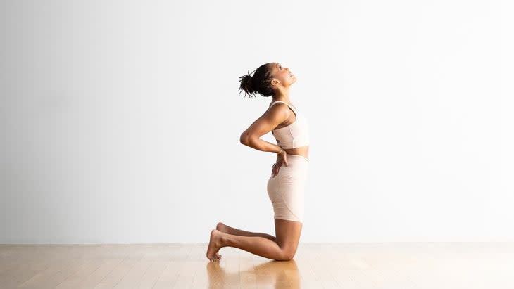 A Black woman with a hair in a loose bun, practices a modified Camel Pose. she is wearing off-white shorts and a cropped top and kneels on a wood floor against a white background. She has her hands on her hips and gazes up at the ceiling.