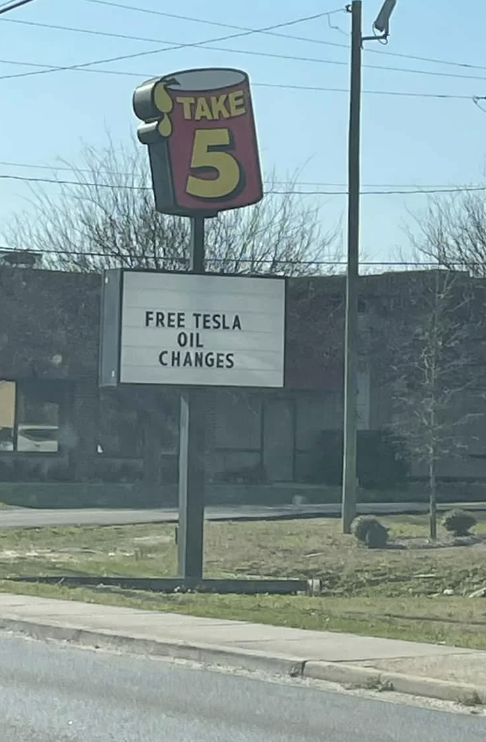 Sign reads "FREE TESLA OIL CHANGES" at a TAKE 5 location, humorously offering a service Teslas do not need