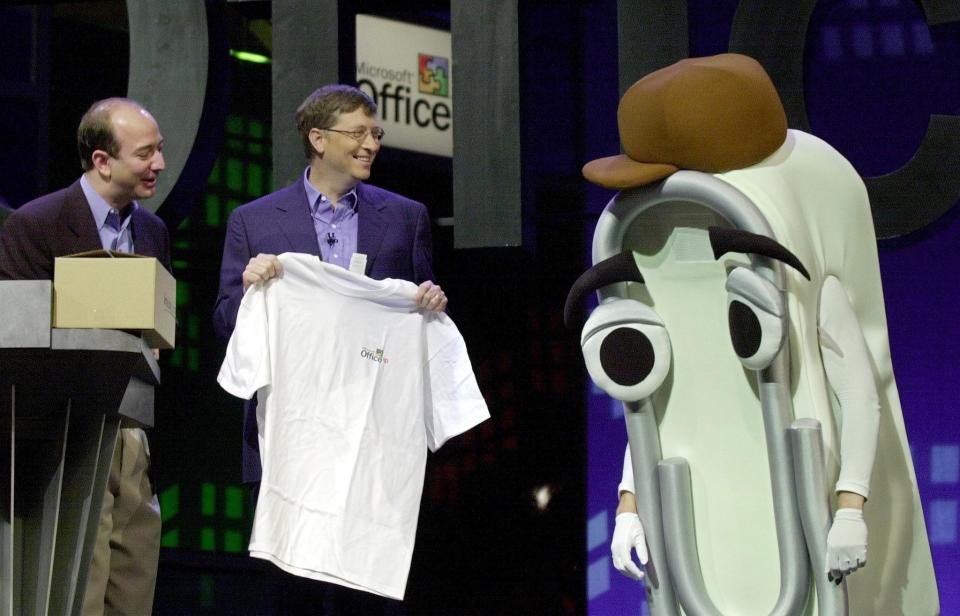 Jeff Bezos, Bill Gates, and a person in a Clippy costume holding a t-shirt at a Microsoft event. The photo features smiles and casual business attire