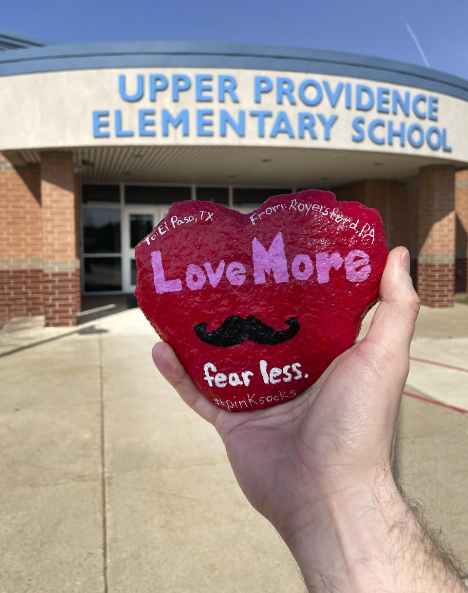 In this March 2021 photo provided by teacher Brian Aikens, Aikens holds up a heart-shaped "kindness rock" memorializing Texas teacher Zelene Blancas, who died of COVID-19 in December 2020, outside Upper Province Elementary School in Royersford, Pa. The stone was decorated and transported across the country by a handful of people to El Paso's Dr. Sue A. Shook Elementary School, where Blancas taught and was remembered by her principal as someone who “embodied kindness.” (Brian Aikens via AP)