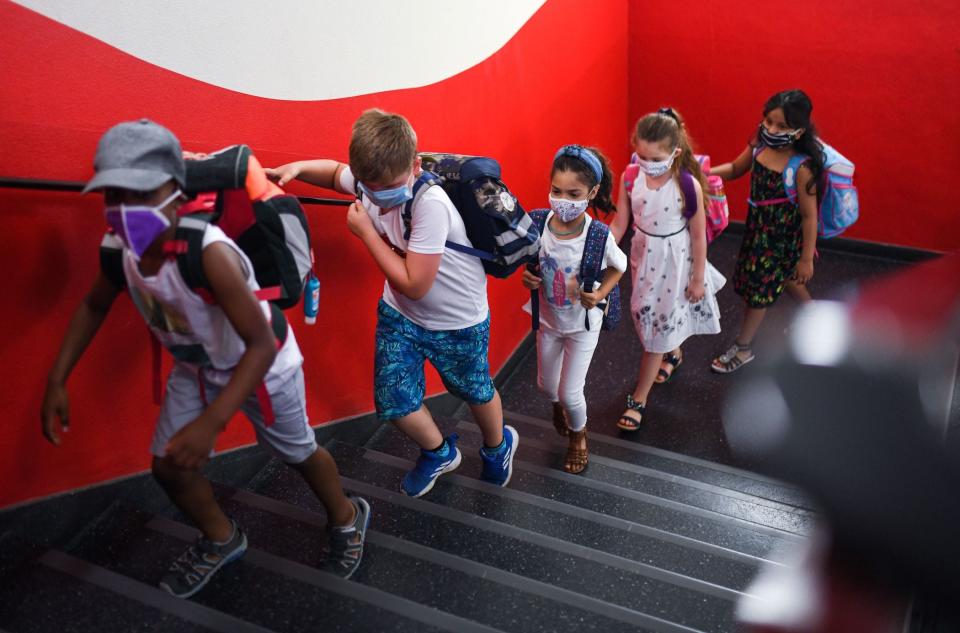 Young kids in masks walk up the stairs against a red wall at a primary school.