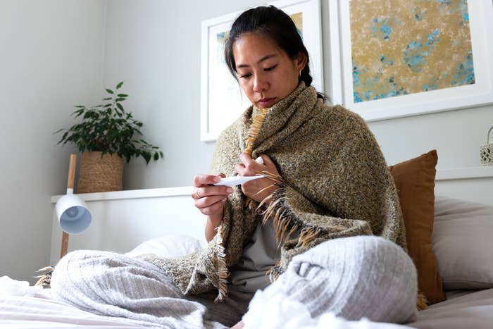 A woman sits on a bed wrapped in a blanket, looking at a thermometer, with framed artwork and a plant in the background