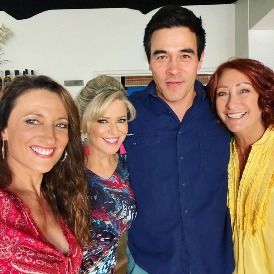 Georgie Parker in a red top, standing next to Emily Symons wearing pink earrings, James Stewart in a blue shirt and Lynne McGranger in a yellow top. They are on the set of Home and Away.