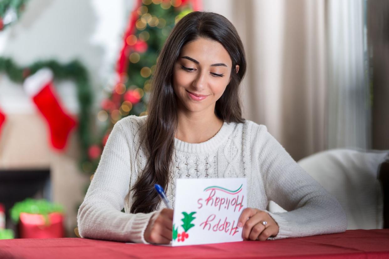 a smiling young woman sits at a table in her decorated living room and looks down in concentration as she writes a letter to a friend for christmas
