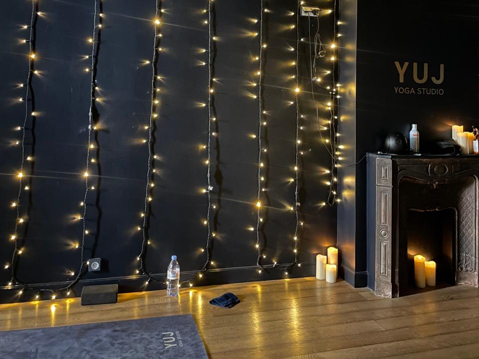 Photo of a wall at the Yuj yoga studio. The wall is painted black and has strands of twinkle lights hanging vertically. On the right is a fireplace with candles sitting inside, on the mantle, and to the left, outside. On the left is a grey yoga mat.