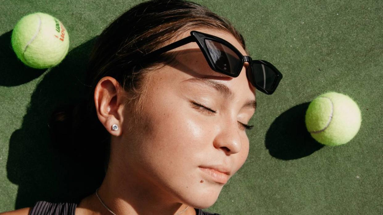  Woman with sunglasses on her head lying down with tennis balls around her. 