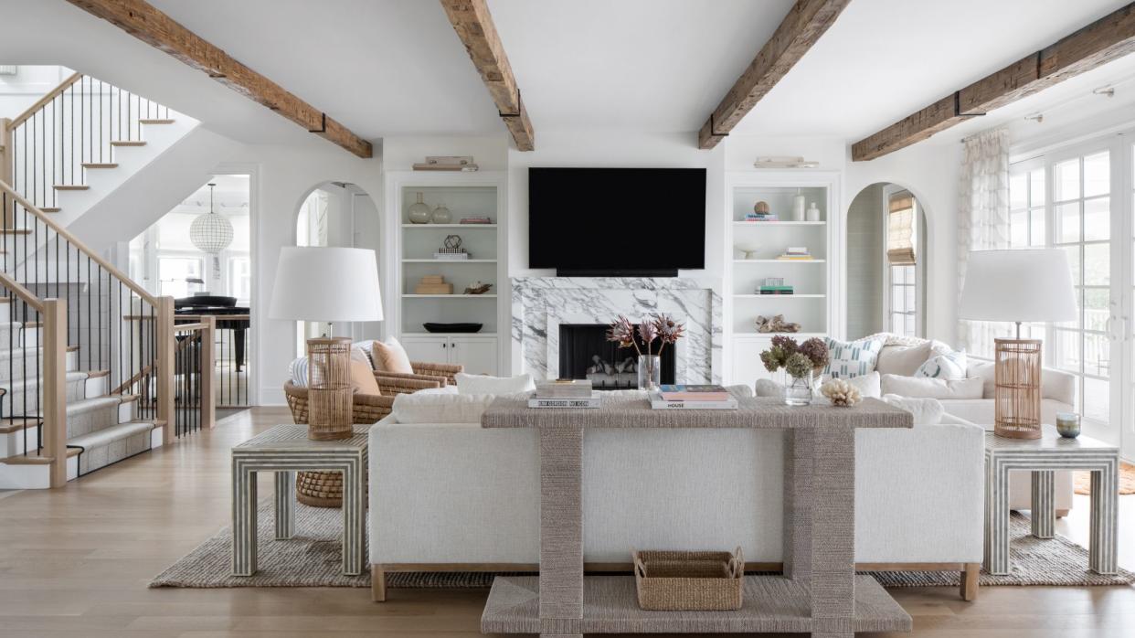  Living room with white walls and white sofa, wood beams in ceiling and TV above marble fireplace. 