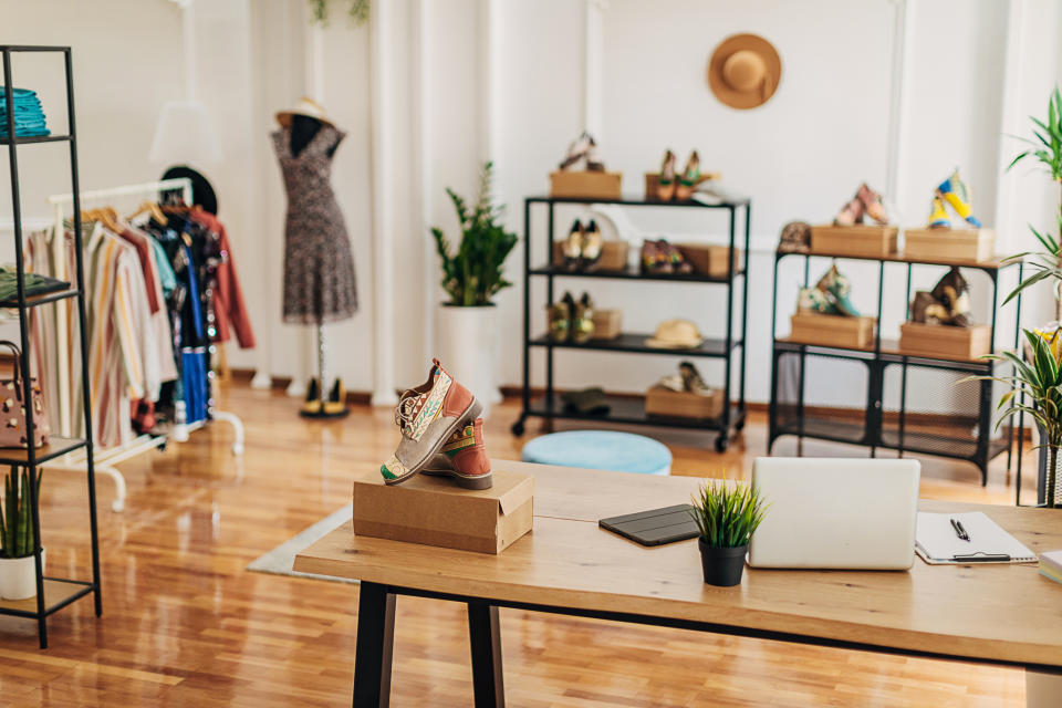 A stylish boutique features a variety of clothing on racks, shoes on shelves, and plants. A desk with a laptop and boxes occupies the foreground