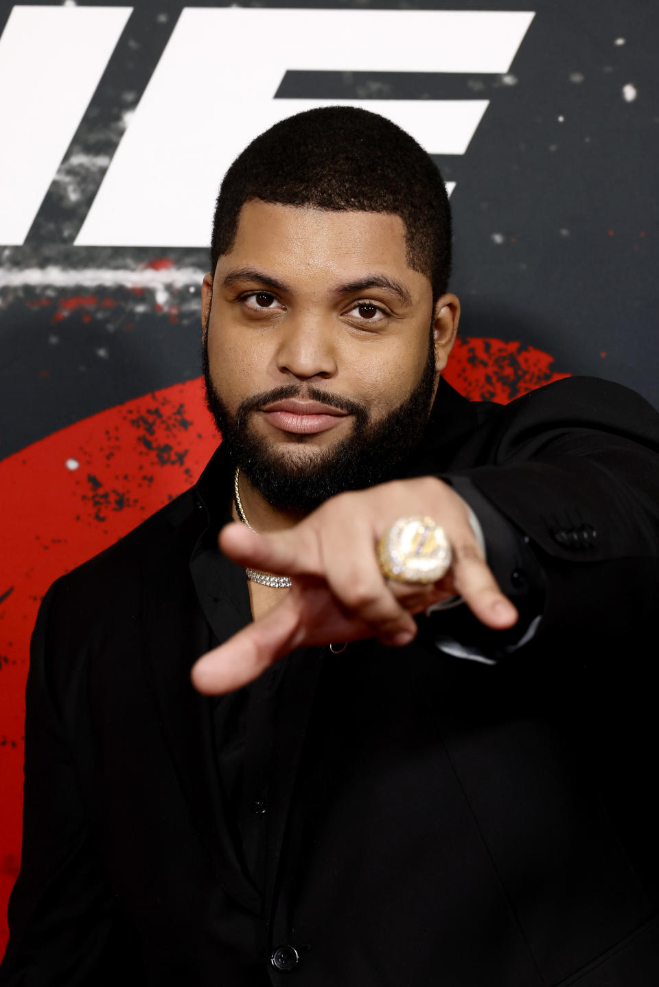 O'Shea Jackson Jr. points towards the camera at an event, wearing a black suit with a gold ring and chain