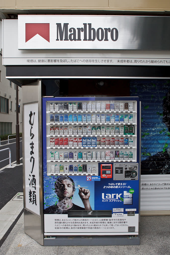 A vending machine in front of a Marlboro store, selling various brands of cigarettes, with advertising featuring a person blowing smoke