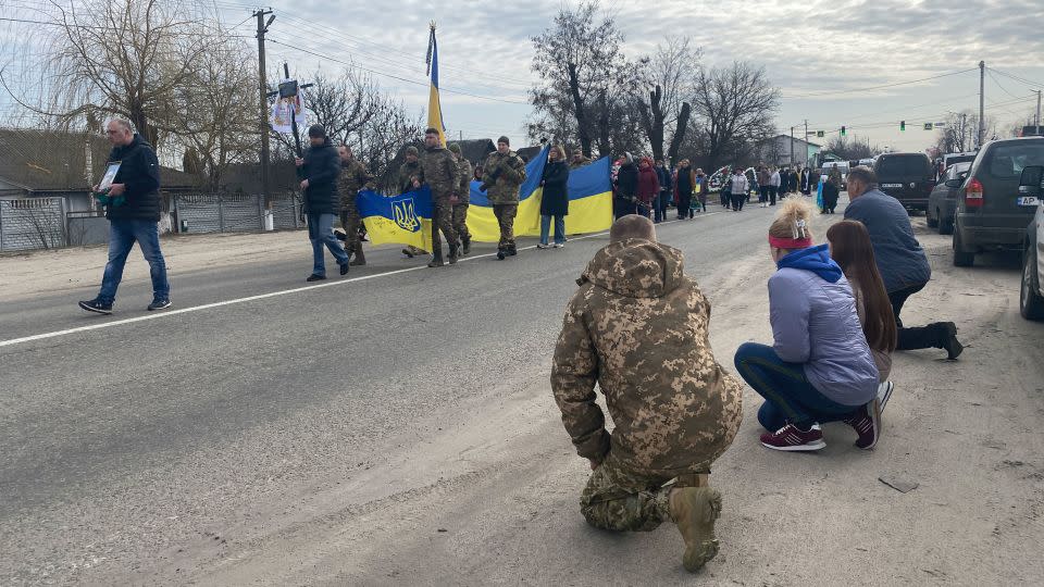 People kneel as they line the main road in the village of Demydiv, just north of Kyiv, while the funeral procession for Oleksandr Savchenko passes by. - Ivana Kottasova/CNN