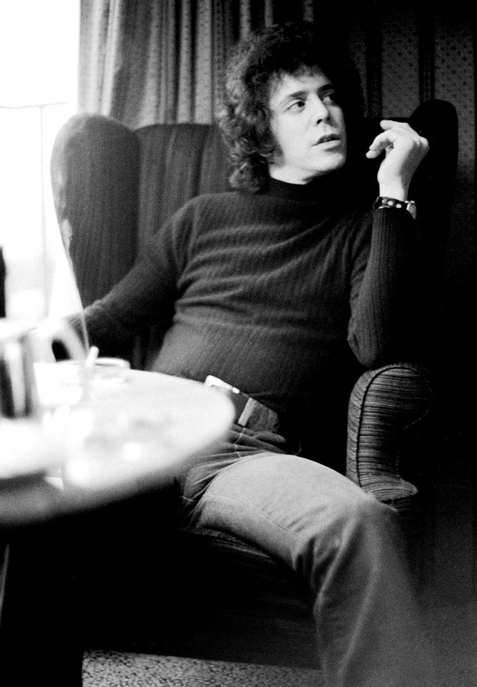 lou reed sits in an armchair and looks to the right, he wears a dark long sleeve shirt and pants with a belt