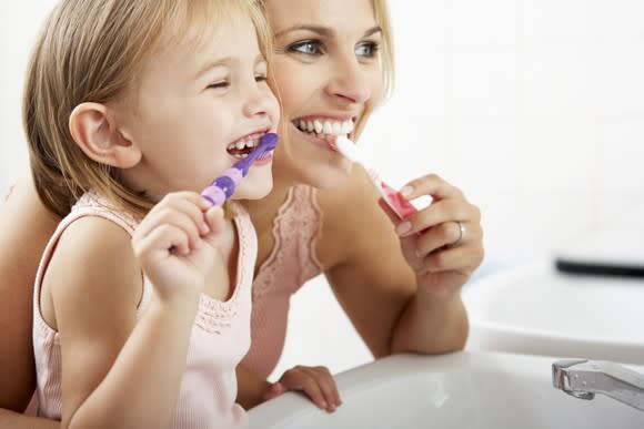 A mom and daughter smile while brushing their teeth.