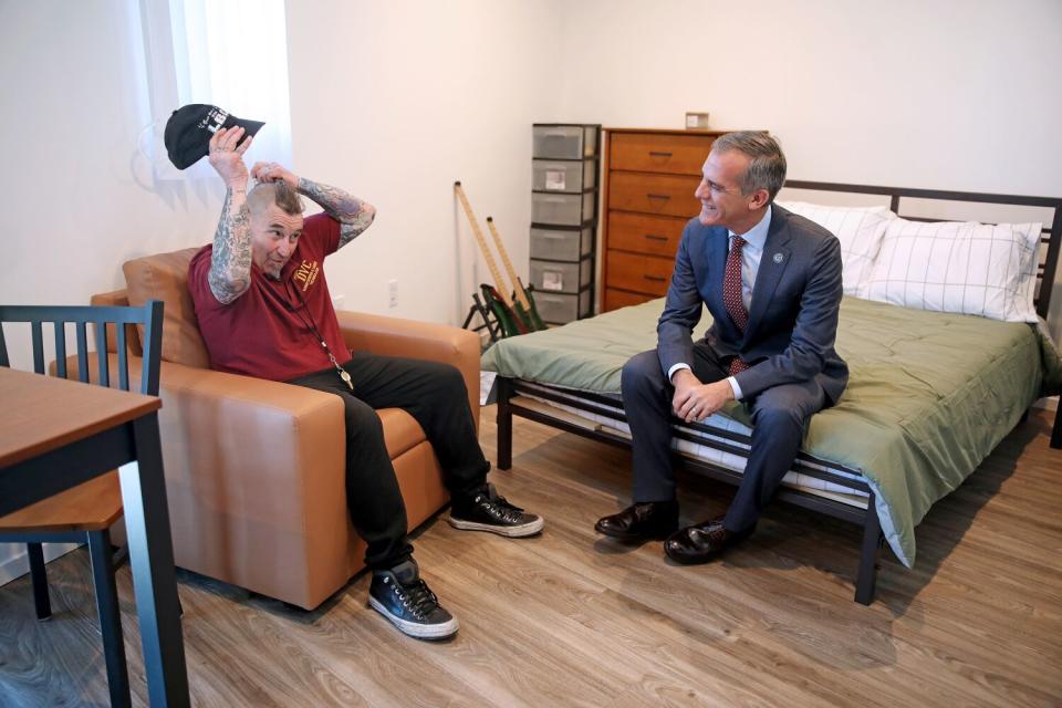 A man in a suit sits on the edge of a bed while talking with a man with a baseball cap sitting in a nearby chair.