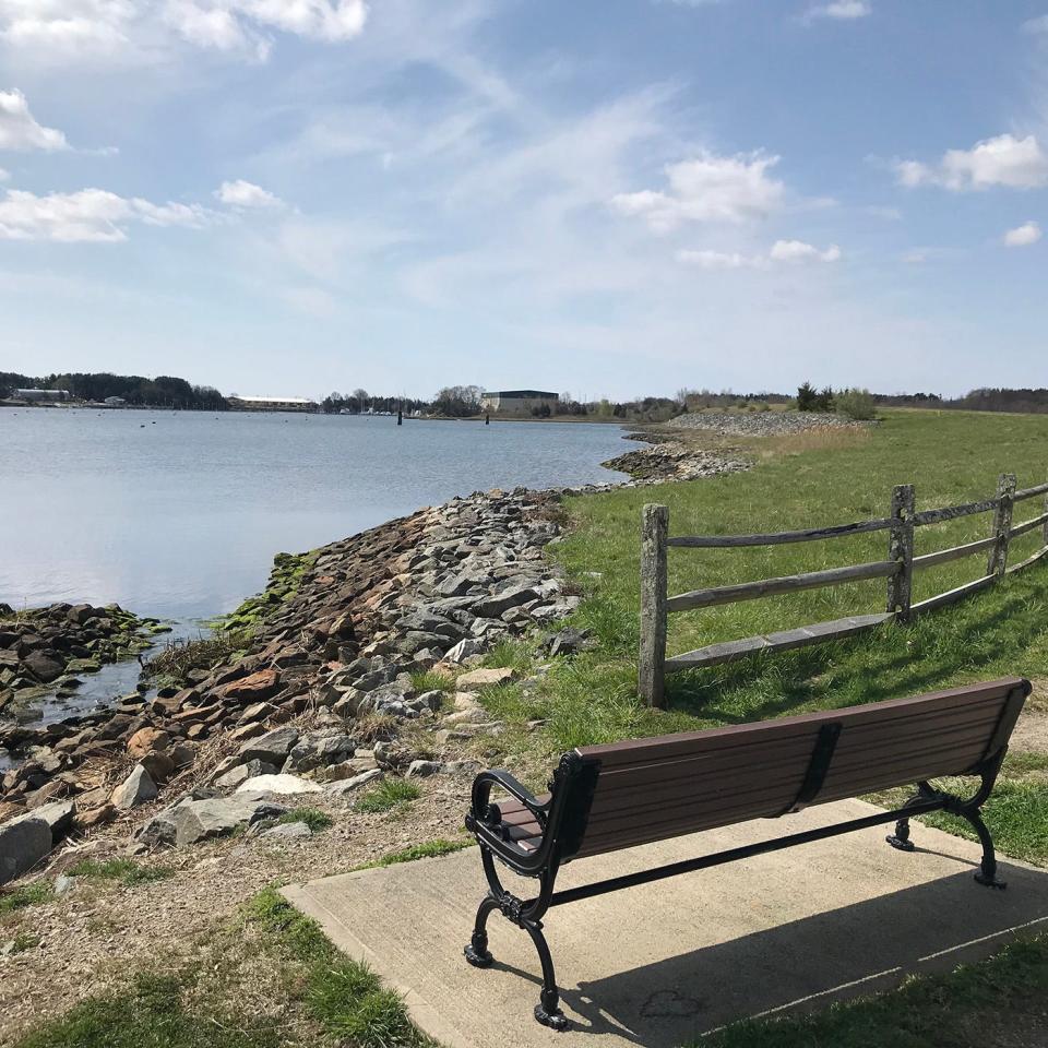 A bench along the bike trail at Calf Pasture Nature Area faces a large field and a stream flowing from Allen Harbor.