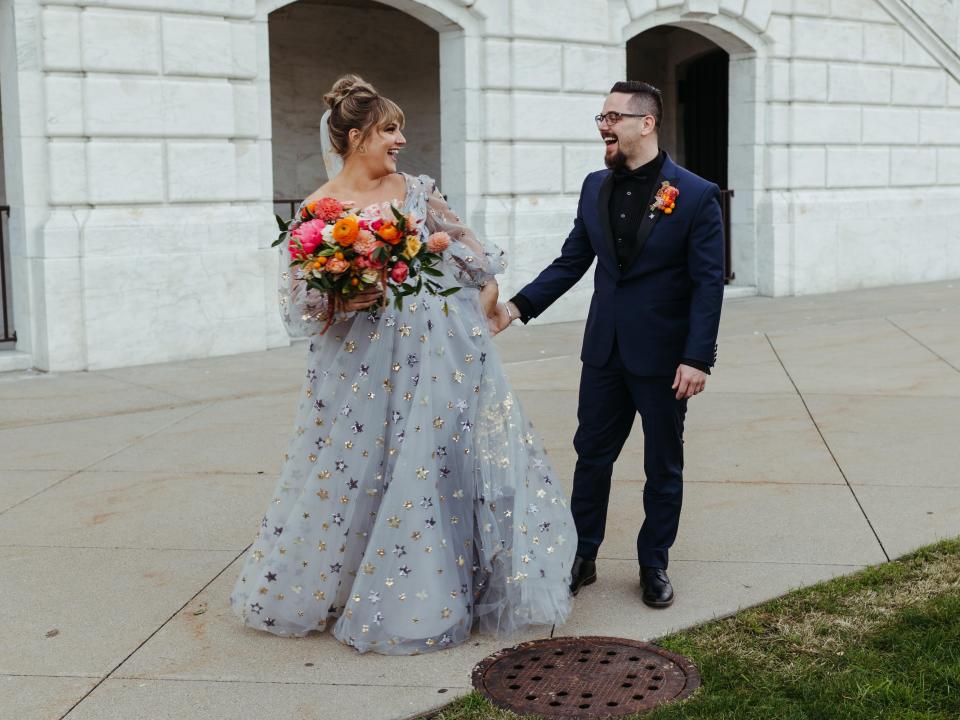 A bride in a blue dress holds hands with her groom in front of a white, stone building.