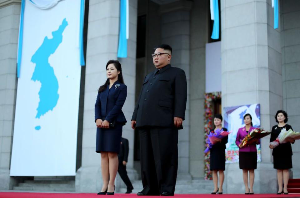 ri sol ju and kim jong un standing on a platform in front of a large white brick building, with banners hanging from the rafters, and three women holding flowers behind them