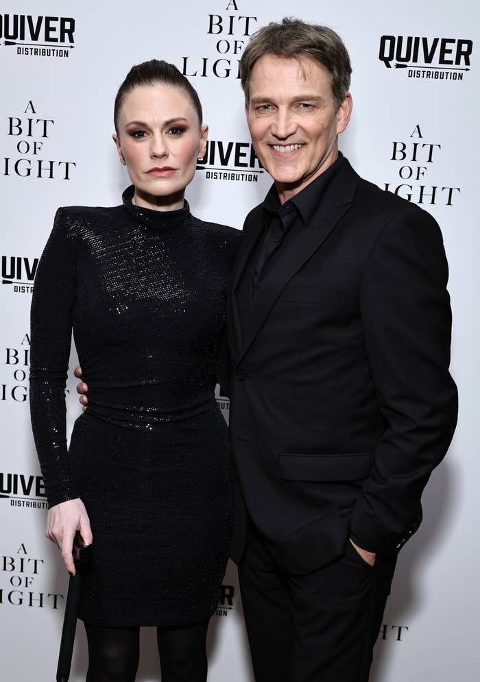 Two people standing side by side; one in a long-sleeve, high-neck dress and the other in a suit with lapels