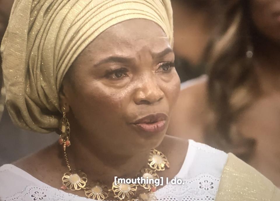 SK's mom in a traditional gele with tears in her eyes mouthing the words I do to encourage SK to say yes to Raven