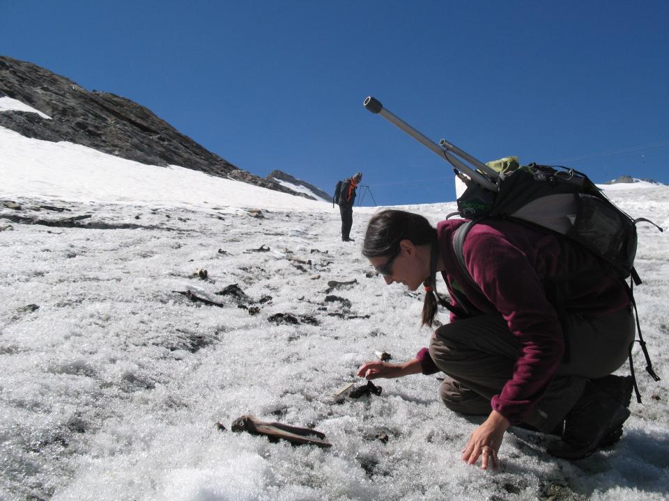 woman wearing large backpack with poles sticking out the top crouches on crunchy textured ice looking at a bone laying on the ground with mountain peaks in the background