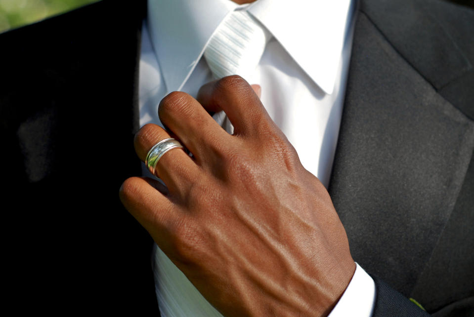 A person adjusts their tie with one hand, showcasing a silver ring on their ring finger. The individual is wearing a suit. Names unknown