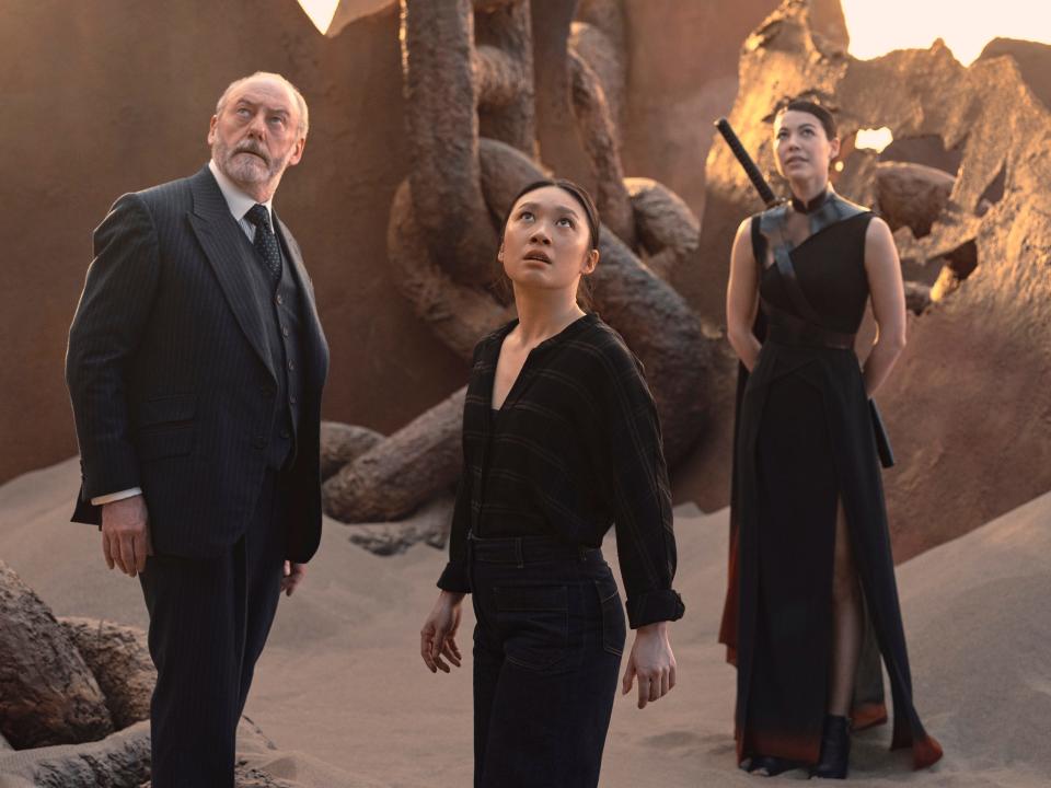 wade, jin cheng, and sophon in 3 body problem, standing in a sandy landscape and looking up at something in the sky. wade is a middle aged man in a suit, jin cheng is a young woman in casual clothing, and sophon is a young woman with a katana strapped to her back and in a flowing dress