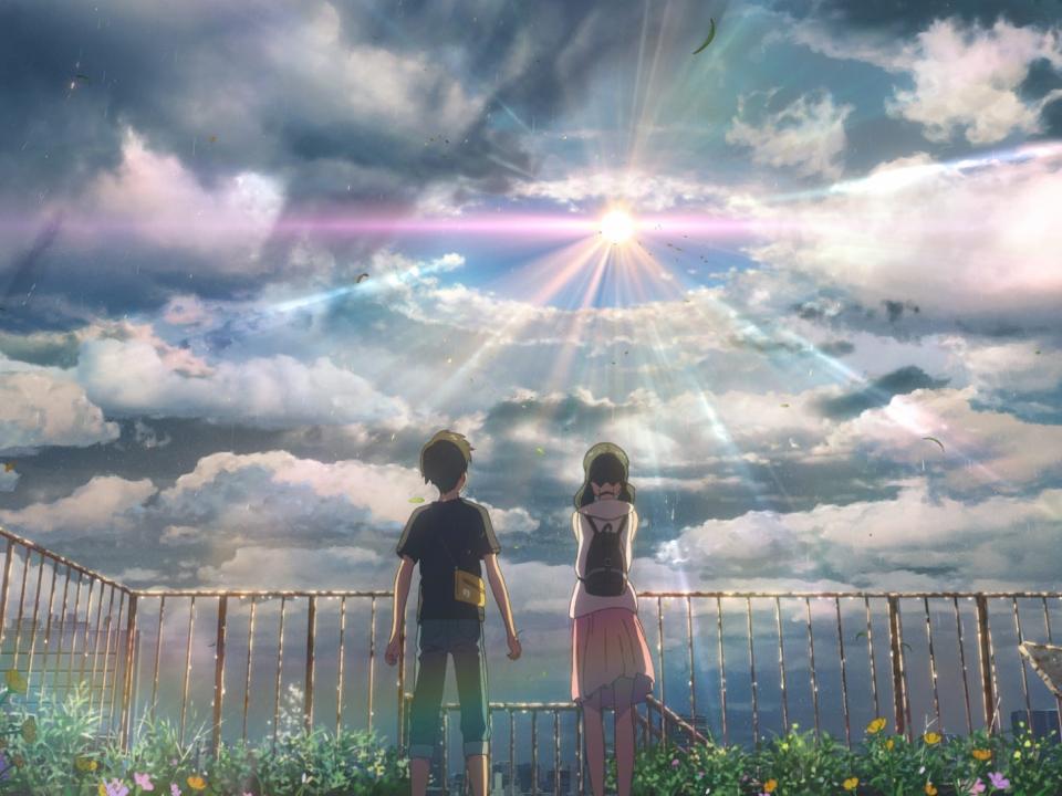 hodaka and hina, a young man and woman, stand on a rooftop in makoto shinkai's weathering with you. their backs are to the viewer as they face the sun, which is brilliantly breaking through an array of clouds. they're surrounded by greenery on the rooftop, which is encircled by railings that seem to be rusted, and leaves flutter in the wind