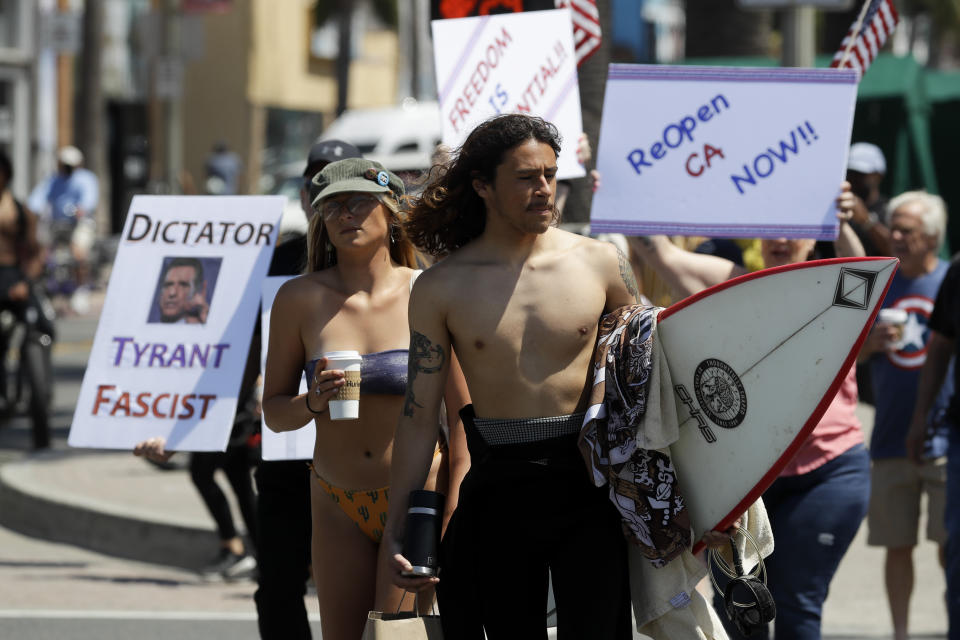 A surfer walks through a crowd of protestors during a May Day demonstration at the pier, Friday, May 1, 2020, in Huntington Beach, Calif. (AP Photo/Chris Carlson)