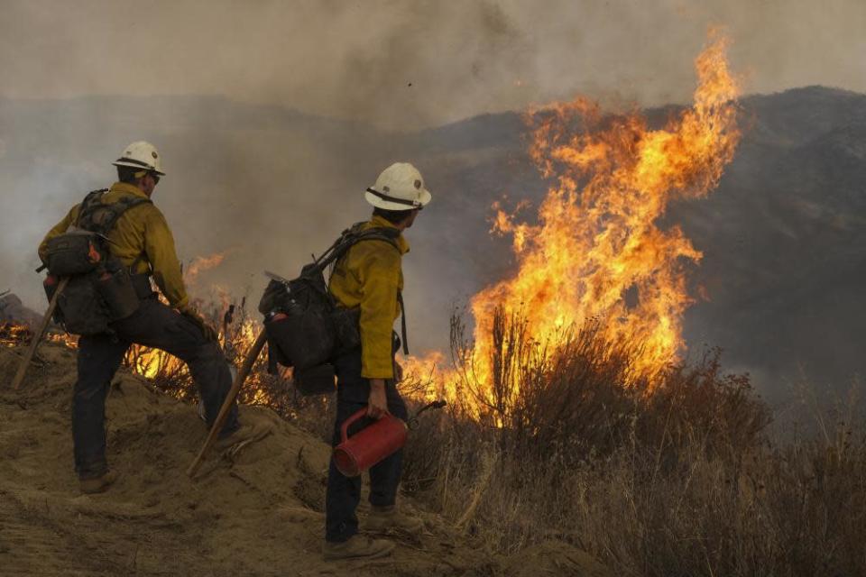 Los bomberos miran hacia la ladera de una colina mientras un fuego arde en la maleza frente a ellos.