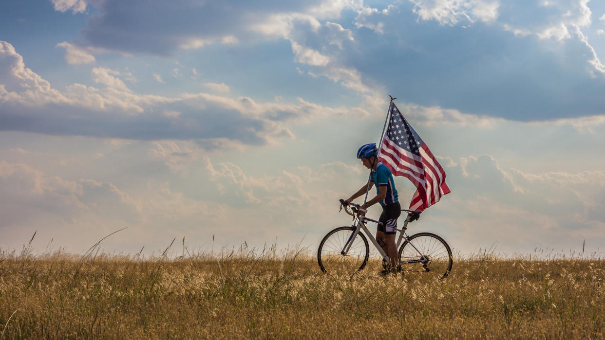  Picture of a cyclist waving american flag in an open field in Oklahoma against a dramatic cloudy sky on Independence Day. 