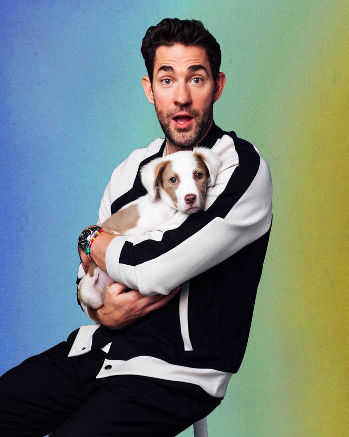John Krasinki  in a striped jacket holding a puppy against a colored backdrop