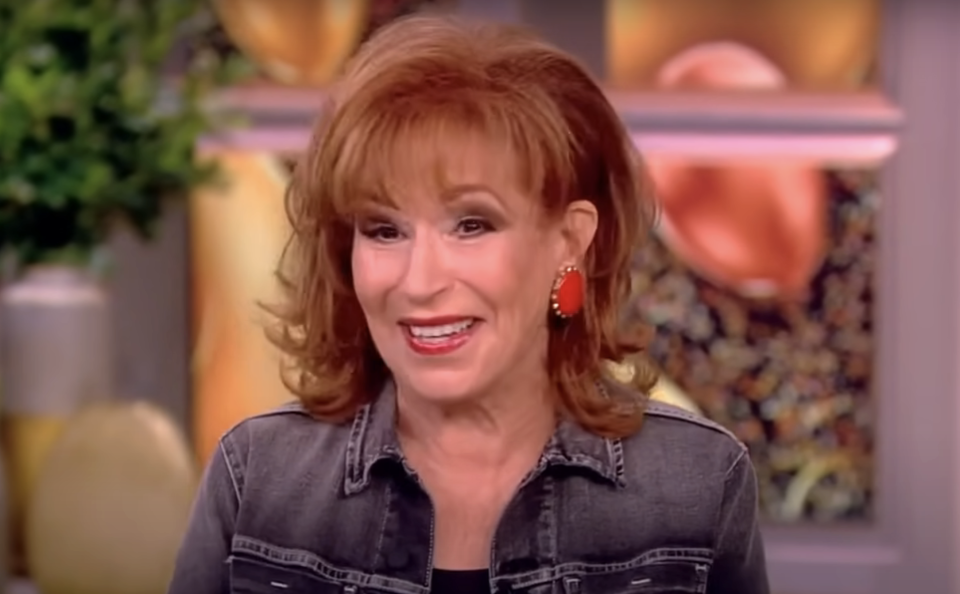 Television personality smiling, wearing denim jacket and red earrings, seated in a studio