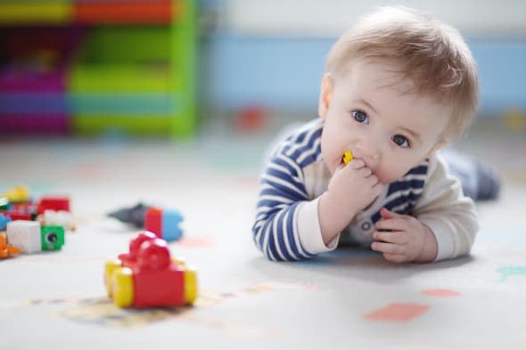 9 month old baby boy lying on his belly, playing with toy bricks, putting one in his mouth