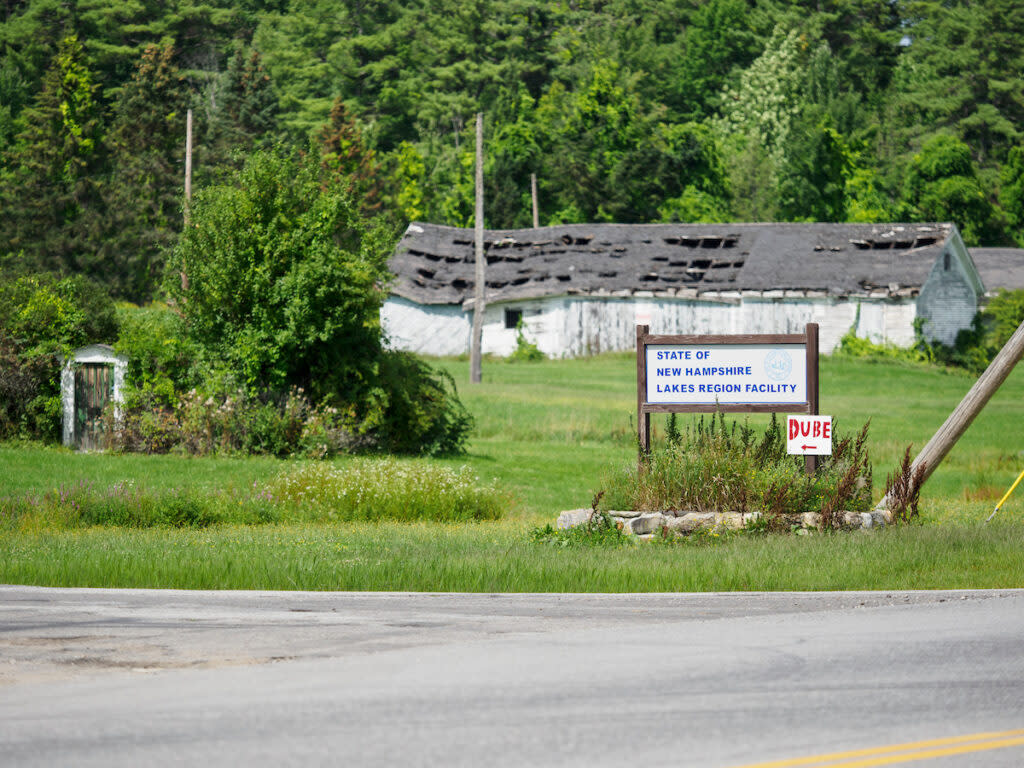 A sign for the Lakes Region Facility, with a decrepit building in the background