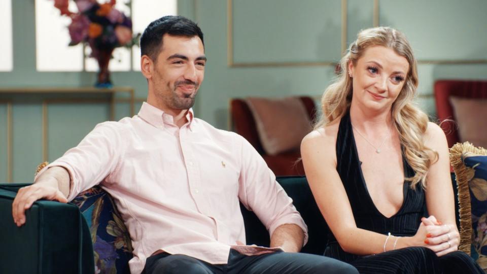 thomas, a man with dark hair and facial stubble, sitting next to rosaline, a woman with long blonde hair wearing a low cut black dress, sitting on a dark green couch and looking at something off camera