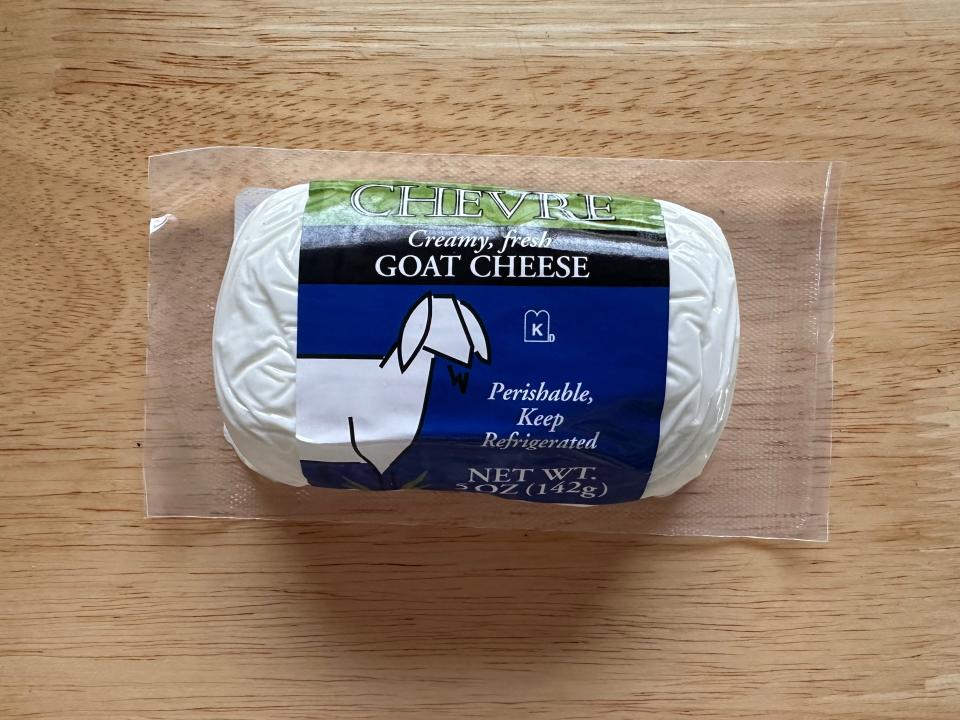 A clear package of goat cheese laying on a wood floor. The label is blue has a graphic of a goat.