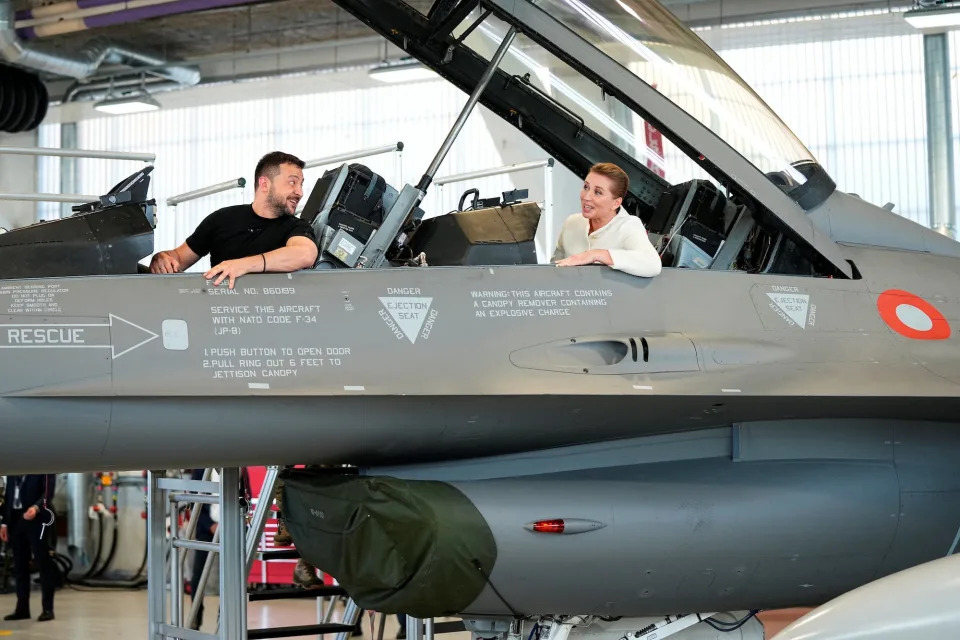 Ukrainian President Volodymyr Zelenskiy sits in front of and  looks back at Denmark's Prime Minister Mette Frederiksen as they sit in a F-16 fighter jet