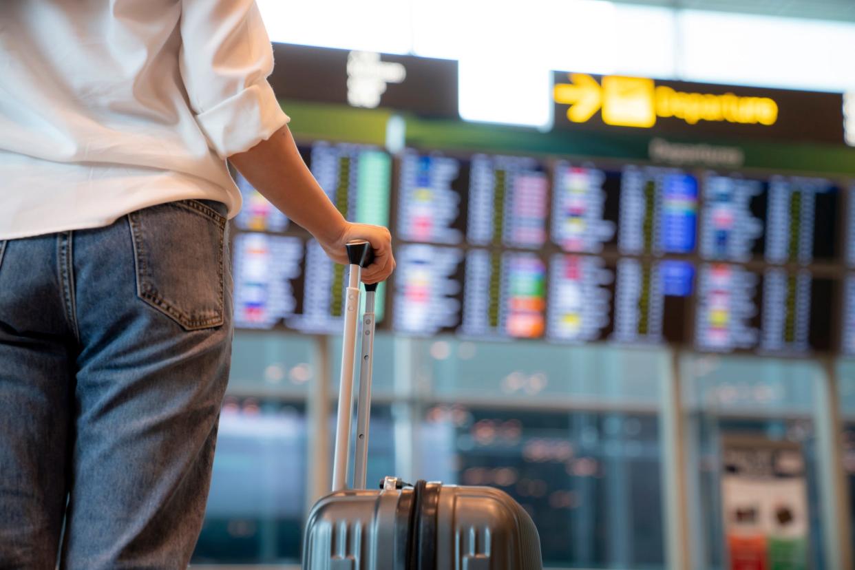 A person holds a suitcase in front of an airport departures board