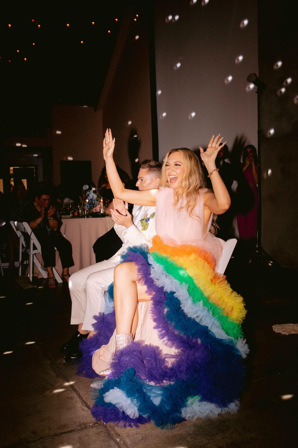 A bride in a rainbow, tulle dress raises her hands in excitement.