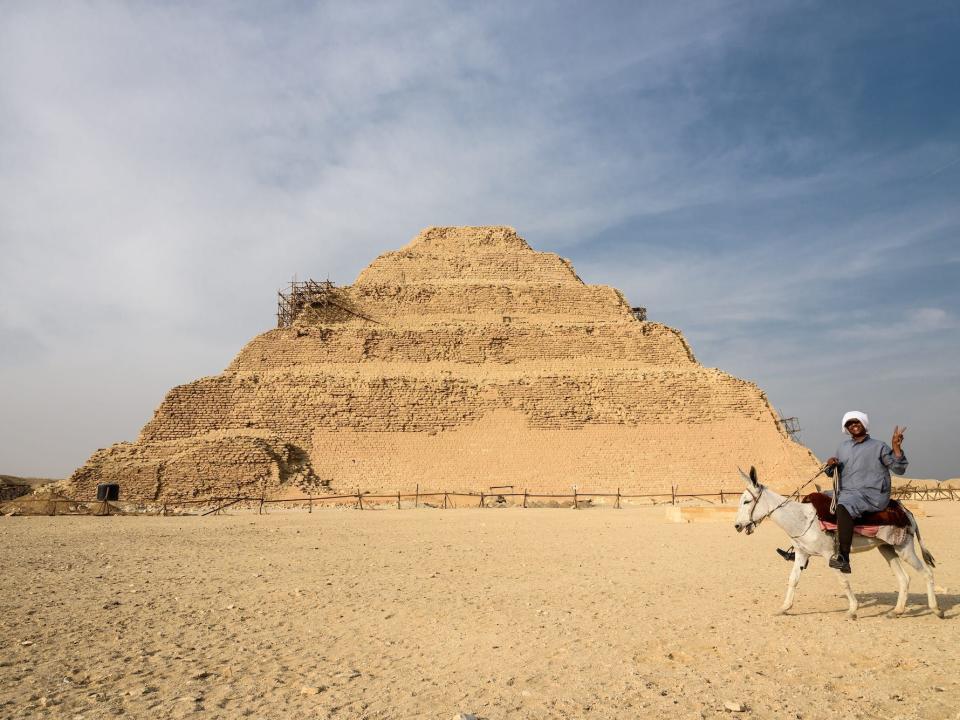 A man rides a donkey, flashing a v for victory sign, in the foreground of this image of the stepped pyramid at Sakkara, Giza, Egypt