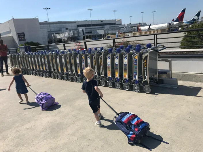 Two children carrying suitcases at an airport outside.