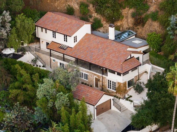 The multi-level home sits perched above the street, surrounded by meticulous landscaping of olive trees, soaring hedges, and lush vegetation.