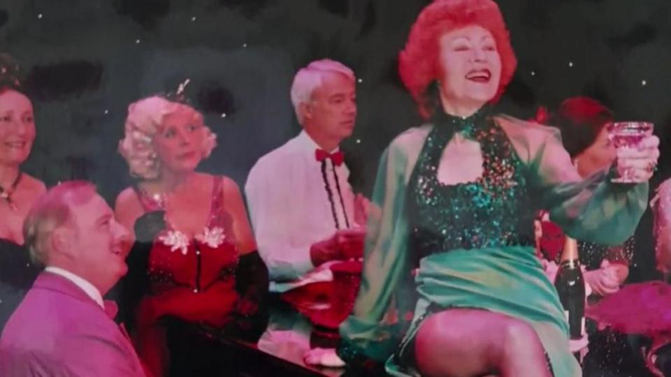 Helen Russell on top of a piano toasting the audience after singing