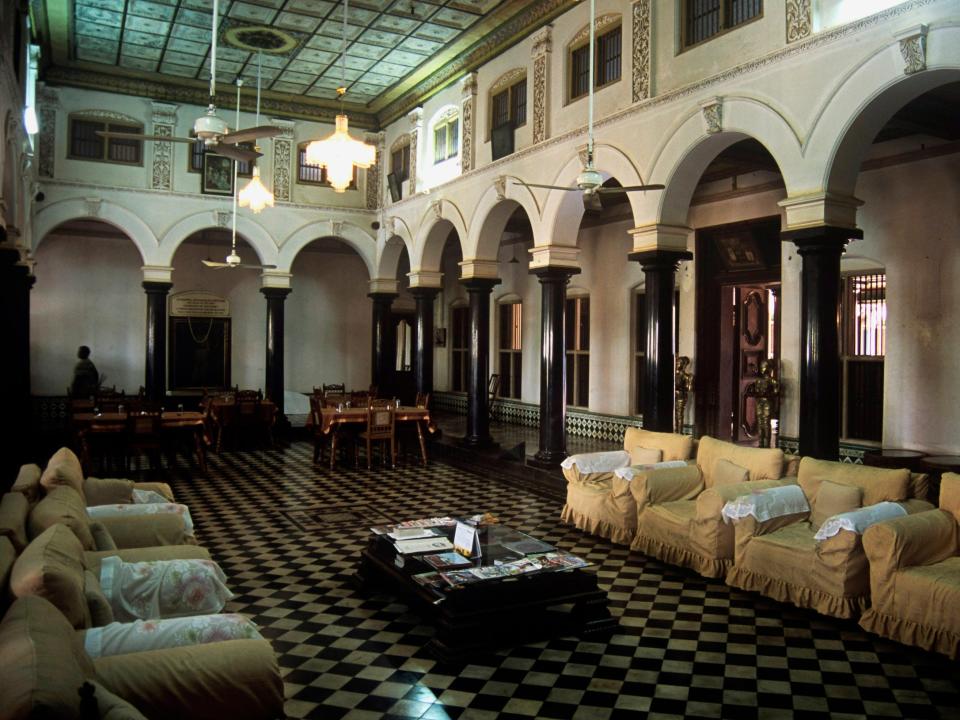 Armchairs, a coffee table, and several dining tables are seen inside a Chettinad mansion.