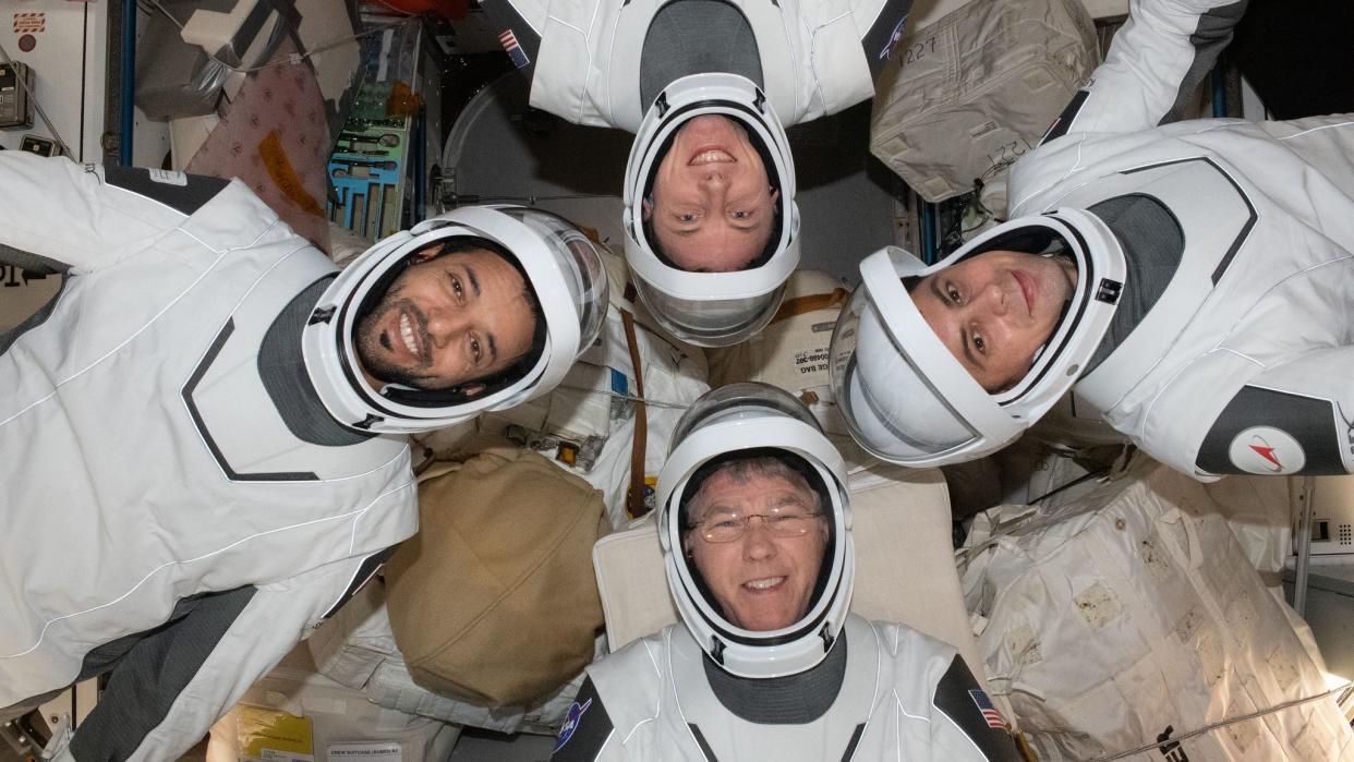  four astronauts in white and gray spacesuits floating in four different directions with their heads pointed to center 