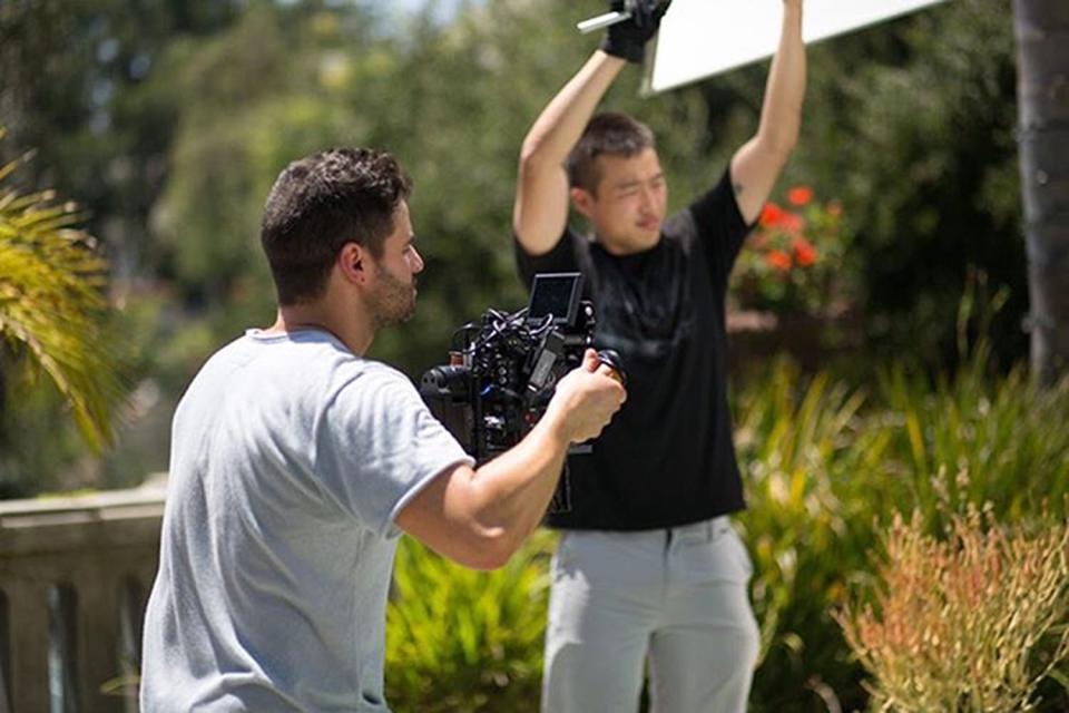 Tim Milgram in a grey T-shirt holding a camera and Isaiah Shinn in a black T-shirt holding lighting on a video shoot.